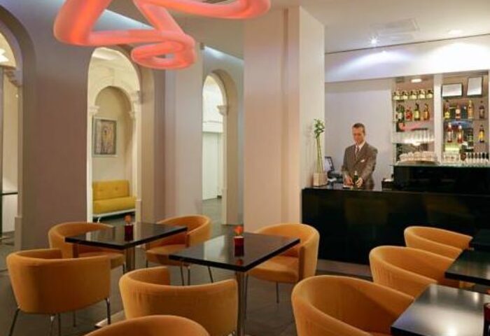 Safestay Vienna gay-friendly hotel bar area. Bartender, tables, orange chairs, and unique lighting. Vienna travel advice for gay men.