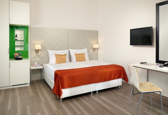 Safestay Vienna gay-friendly hotel room. Double bed with orange throw, desk, TV, and storage unit. Modern, clean design for gay travelers.