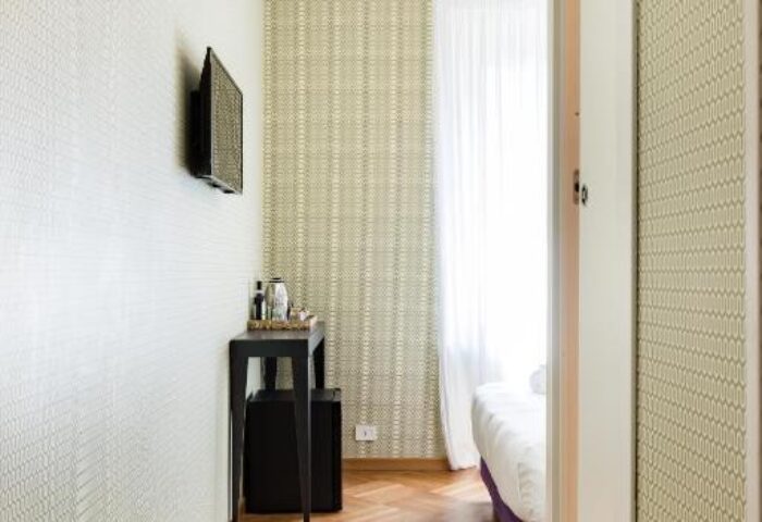 Salotto Monti gay-friendly hotel room. View of bed, herringbone wood floor, patterned wallpaper, TV, small table, and mini fridge.