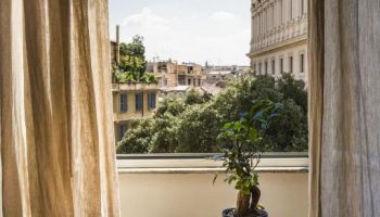 Salotto Monti gay-friendly hotel room view. Table with books and plant. Balcony overlooking Rome's cityscape. Interior design, travel advice.
