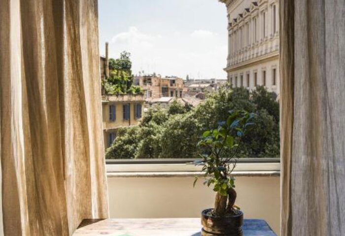 Salotto Monti gay-friendly hotel room view. Table with books and plant. Balcony overlooking Rome's cityscape. Interior design, travel advice.