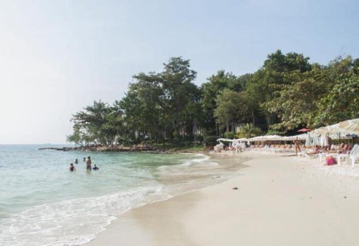 Beach at Samed Villa Resort, a gay-friendly hotel. People swim in the sea, others relax on beach chairs under umbrellas. Tropical trees backdrop.