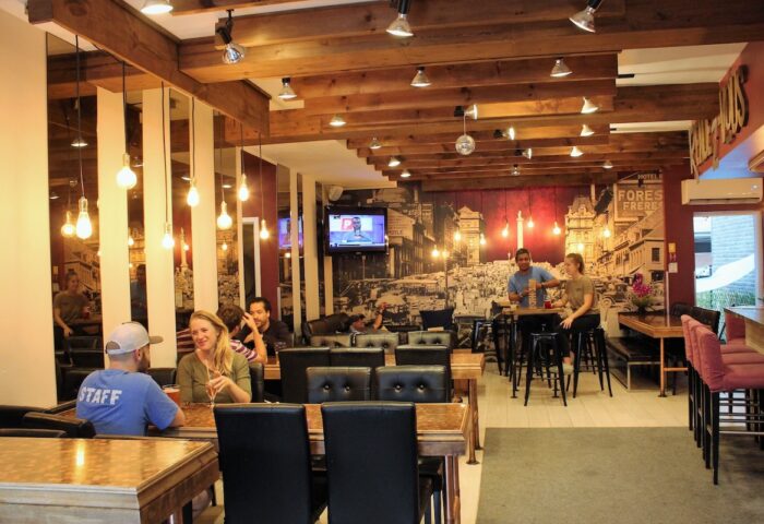 Samesun Montreal Central gay-friendly hotel interior. Guests at tables, bar area, and a vintage city mural. Wooden beams and warm lighting.