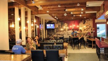 Samesun Montreal Central gay-friendly hotel interior. Guests at tables, bar area, and a vintage city mural. Wooden beams and warm lighting.