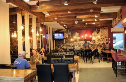 Samesun Montreal Central gay-friendly hotel interior. Guests at tables, bar area, and a vintage city mural. Wooden beams and warm lighting.
