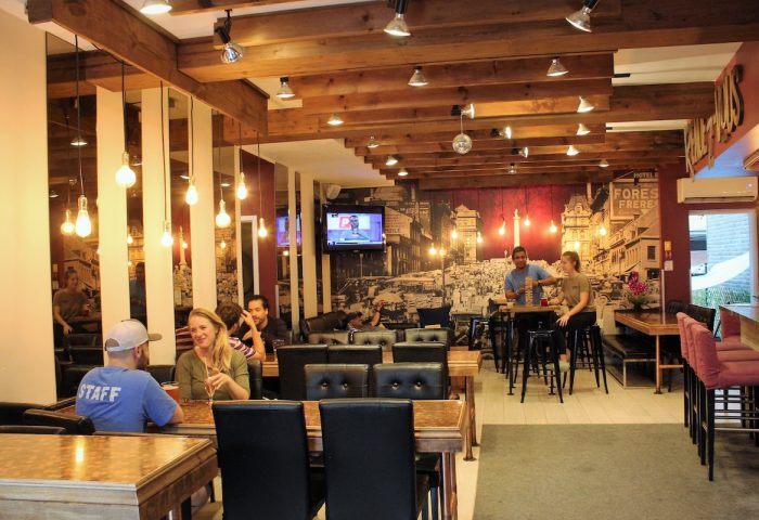 Samesun Montreal Central gay-friendly hotel interior. Guests at tables, bar area, and a vintage city mural. Wooden beams and warm lighting.