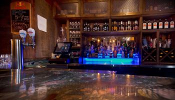 Bar at Samesun Montreal Central, a gay-friendly hotel. Shelves of liquor bottles, glasses, and a blue-lit bar top. Montreal travel.