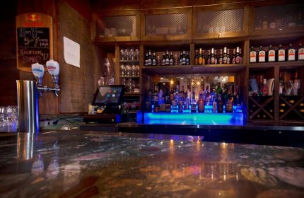 Bar at Samesun Montreal Central, a gay-friendly hotel. Shelves of liquor bottles, glasses, and a blue-lit bar top. Montreal travel.