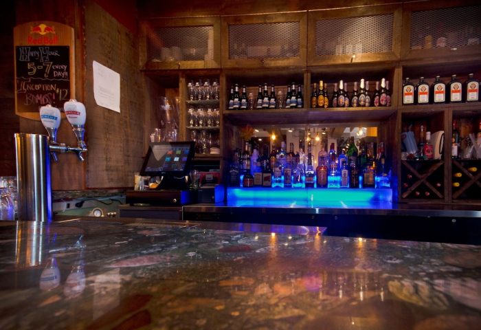 Bar at Samesun Montreal Central, a gay-friendly hotel. Shelves of liquor bottles, glasses, and a blue-lit bar top. Montreal travel.