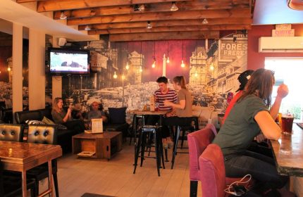 Samesun Montreal Central's gay-friendly hotel bar area. People sit at tables, bar, and couches. Vintage Montreal mural on the wall.