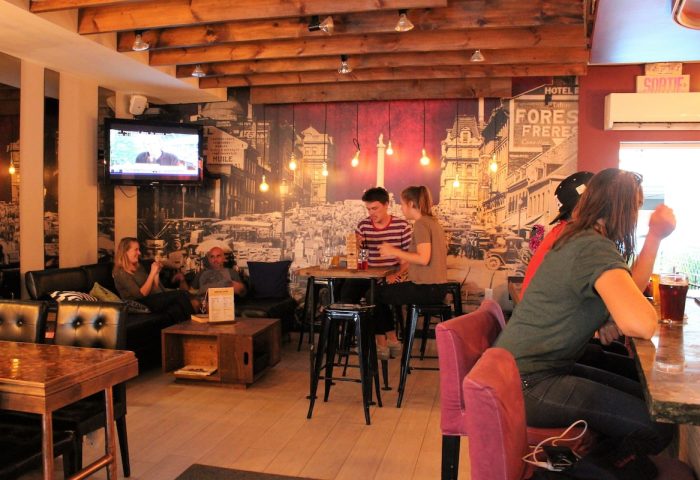 Samesun Montreal Central's gay-friendly hotel bar area. People sit at tables, bar, and couches. Vintage Montreal mural on the wall.