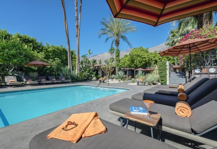 Poolside view at Santiago Resort, a gay-friendly hotel in Palm Springs. Loungers, cocktail, towel, and lush greenery create a relaxing atmosphere.