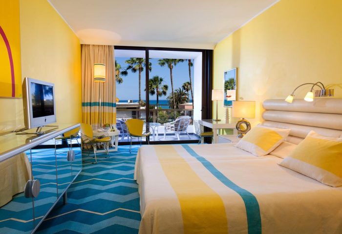 Seaside Palm Beach gay-friendly hotel room. Balcony view of palm trees and ocean. Bright yellow decor, blue carpet, and modern furniture.