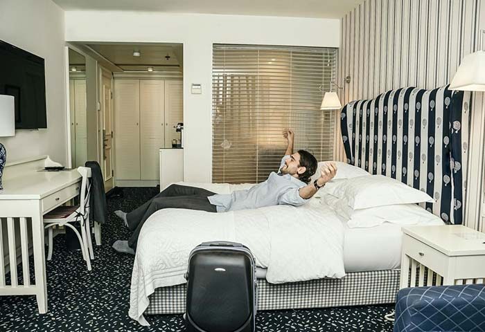 Gay-friendly Shalom Hotel & Relax, an Atlas Boutique Hotel. Man relaxing on bed in pet-friendly room. Suitcase. Gay men travel advice.