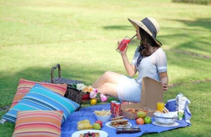 Picnic at Shangri-la Hotel Jakarta, a gay-friendly hotel. Woman with hat drinks juice. Food, basket, pillows on blue blanket in green grass.