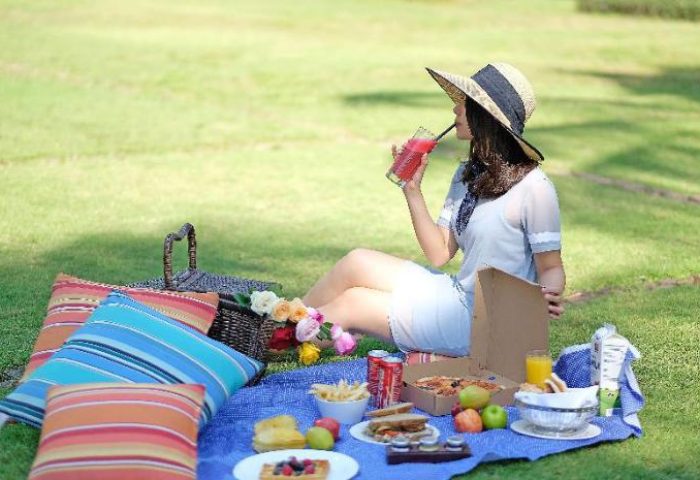 Picnic at Shangri-la Hotel Jakarta, a gay-friendly hotel. Woman with hat drinks juice. Food, basket, pillows on blue blanket in green grass.