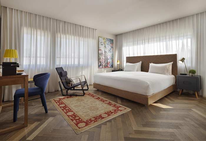 Shenkin Hotel room: Modern gay-friendly hotel bedroom with bed, chair, desk, herringbone wood floor, and light filtering curtains. Interior design.