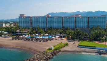 Gay-friendly Sheraton Buganvilias Resort and Convention Center aerial view. Beachfront hotel with pools, palm trees, and mountains in the background.