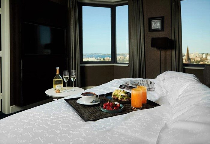 Sheraton Grand Sydney Hyde Park gay-friendly hotel room. Breakfast in bed with city view. Champagne, juice, coffee, fruit, and avocado toast.