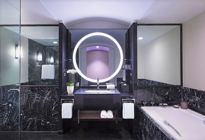 Sheraton Grand Sydney Hyde Park gay-friendly hotel bathroom. Circular mirror, marble walls, towels, and bathtub. Luxury design.