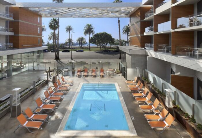 Shore Hotel's pool area, a gay-friendly hotel. Palm trees and ocean views. Lounge chairs surround the pool. Modern architecture.