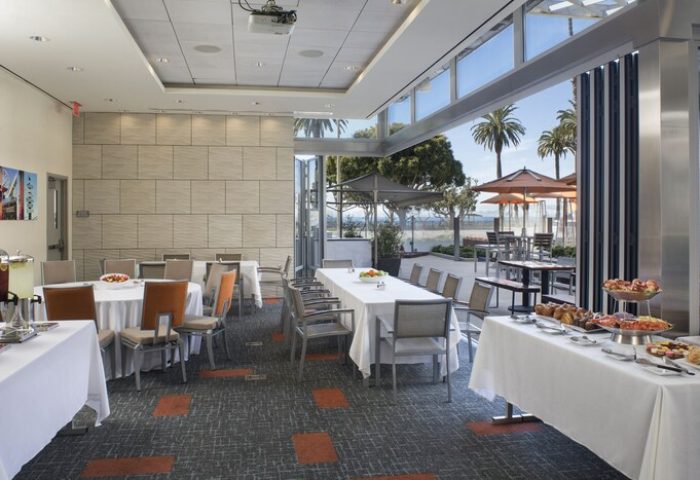 Shore Hotel's event space with tables, chairs, and buffet. Open doors lead to an outdoor patio with palm trees. Gay-friendly hotel.