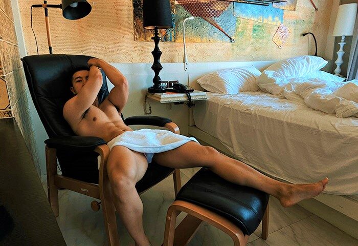 Gay-friendly Siam @ Siam Design Hotel Bangkok: A shirtless man lounges in a chair, towel-clad, in a stylish room. Gay travel luxury.