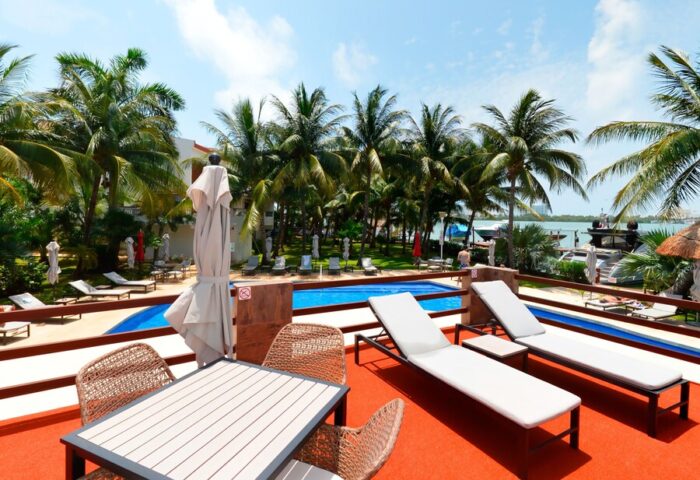 Sina Suites gay-friendly hotel pool view. Loungers and table with chairs on deck. Palm trees, blue sky, and ocean backdrop.