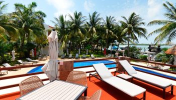 Sina Suites gay-friendly hotel pool view. Loungers and table with chairs on deck. Palm trees, blue sky, and ocean backdrop.