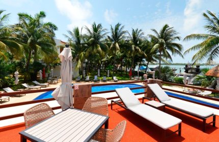 Sina Suites gay-friendly hotel pool view. Loungers and table with chairs on deck. Palm trees, blue sky, and ocean backdrop.