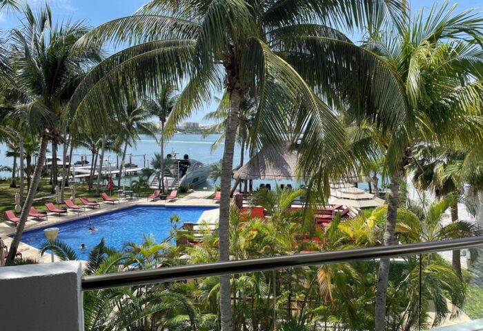 Sina Suites gay-friendly hotel view. Palm trees frame a pool with lounge chairs, a palapa, and ocean views. A relaxing tropical gay vacation spot.