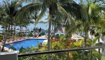 Sina Suites gay-friendly hotel view. Palm trees frame a pool with lounge chairs, a palapa, and ocean views. A relaxing tropical gay vacation spot.