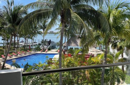 Sina Suites gay-friendly hotel view. Palm trees frame a pool with lounge chairs, a palapa, and ocean views. A relaxing tropical gay vacation spot.