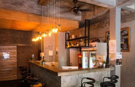 Bar area at The Beach Hostel, a gay-friendly hotel. Pendant lights hang above the counter with bar stools. Shelves of liquor behind.