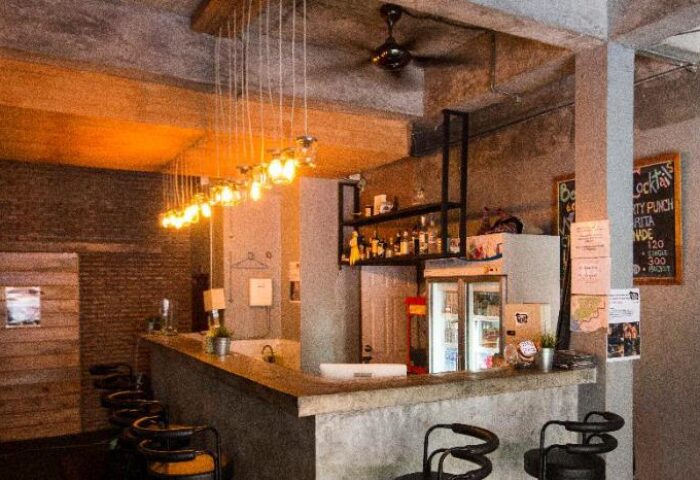Bar area at The Beach Hostel, a gay-friendly hotel. Pendant lights hang above the counter with bar stools. Shelves of liquor behind.