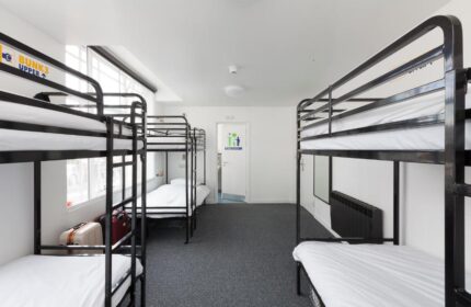 So Hostel gay-friendly hotel dorm room. Bunk beds with white sheets, gray carpet, & bathroom door. Suitcases under a bed. Clean, modern hostel.