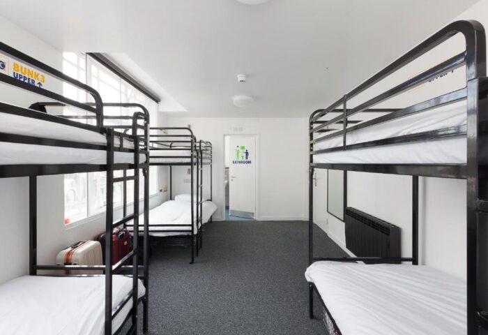 So Hostel gay-friendly hotel dorm room. Bunk beds with white sheets, gray carpet, & bathroom door. Suitcases under a bed. Clean, modern hostel.
