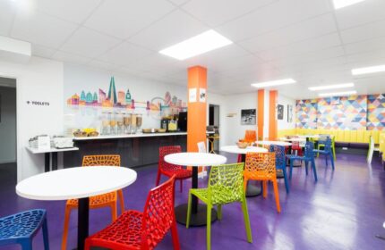 So Hostel's dining area, a gay-friendly hotel. Colorful chairs and tables on a purple floor. London skyline mural. Breakfast buffet.