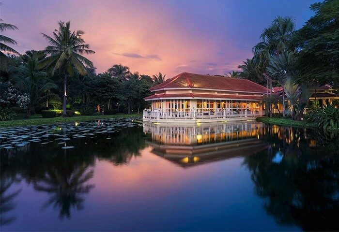 Sofitel Angkor Phokeethra Golf & Spa Resort view at dusk. Gay-friendly hotel with building reflected in water, palm trees, and colorful sky.