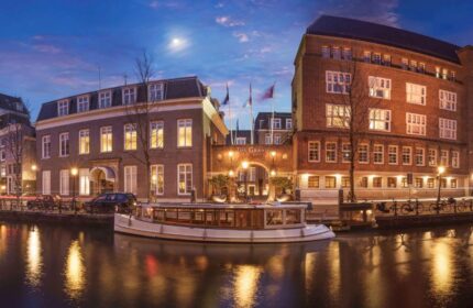 Sofitel Legend The Grand Amsterdam, a gay-friendly hotel, at dusk. Canal boat reflects lights. Elegant architecture, a top choice for gay travel.