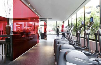Sofitel Los Angeles at Beverly Hills Hotel gym. Gay-friendly hotel with modern fitness equipment, red glass wall, and natural light. Workout space.