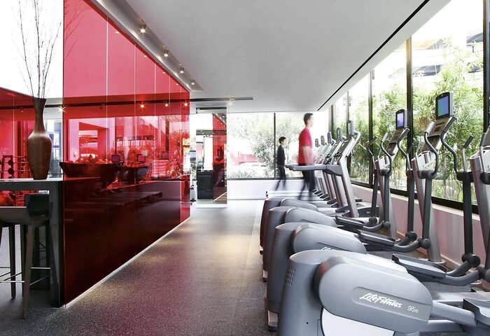 Sofitel Los Angeles at Beverly Hills Hotel gym. Gay-friendly hotel with modern fitness equipment, red glass wall, and natural light. Workout space.