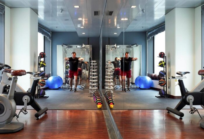Gym at Sofitel Munich Bayerpost Hotel. Man exercising in the gay-friendly hotel's fitness center with weights, exercise bike, and mirrored wall.