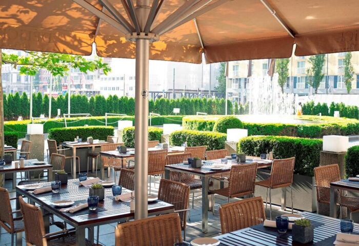 Sofitel Munich Bayerpost Hotel outdoor dining. Gay-friendly hotel with tables, chairs, and umbrellas. Fountain and greenery in the background.
