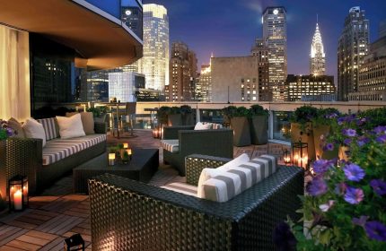 Sofitel New York gay-friendly hotel rooftop lounge at night. City skyline view, stylish seating, and flowers. Luxury travel for gay men.