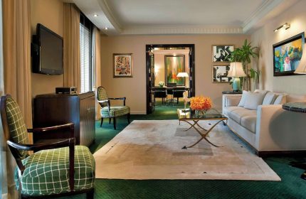 Sofitel New York suite living room. Gay-friendly hotel with sofa, chairs, TV, and desk visible through doorway. Upscale travel for gay men.