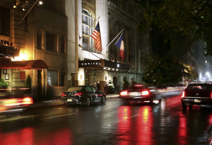 Night view of the Sofitel New York, a gay-friendly hotel. Flags of the USA and France fly above the entrance. Cars drive on wet street.
