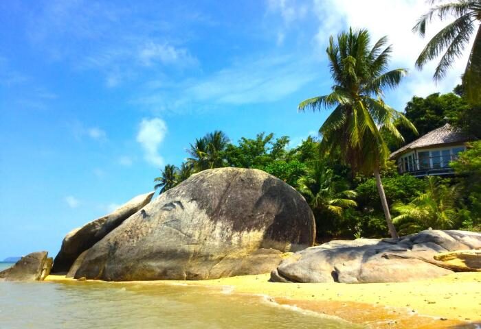Gay-friendly hotel, Somewhere Only We Know. Beach view with rocks, palm trees, and a building in the background. Tropical paradise for gay travel.