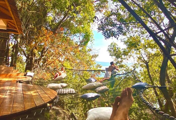 Gay-friendly hotel Somewhere Only We Know: Men relax on a net strung between trees, pillows scattered around. Tropical gay travel destination.