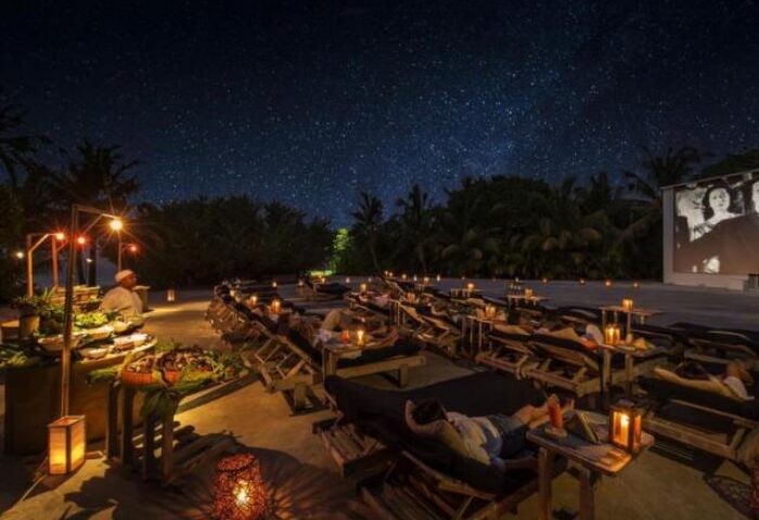 Outdoor cinema at Soneva Fushi, a gay-friendly hotel. Guests relax on loungers under the stars, enjoying a movie and buffet.